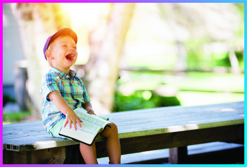 Boy in shorts and plaid shirt sitting on a table, laughing, while reading the Holy Bible.