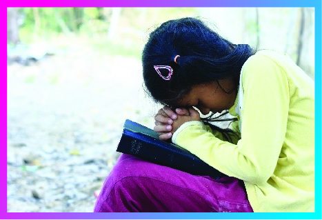 Girl in yellow shirt and pink pants prays, with her head and hands resting on the Holy Bible.